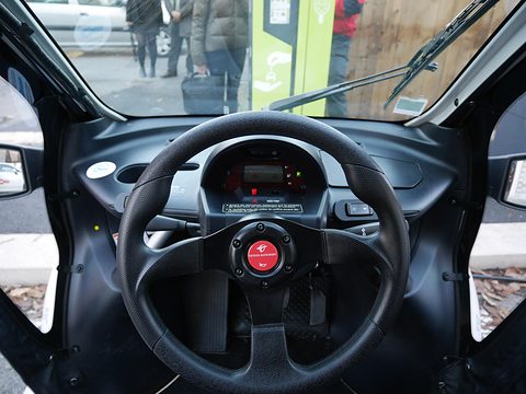 Black three-spoke steering wheel in front of a small digital display, seen through the open side of the car. Opens a larger view