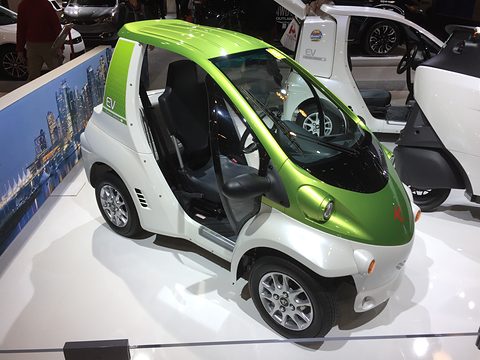 Green and white single-seat microcar with an open side on a white stand, a city-scene backdrop behind. Opens a larger view