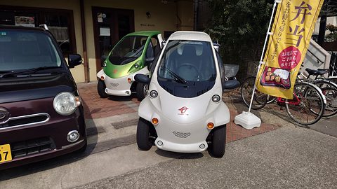 A white and a green single-seat microcar parked by a shop banner, a dark kei hatchback alongside. Opens a larger view