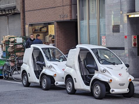 Two white single-seat microcars with clear doors parked nose to tail outside a shop with bicycles. Opens a larger view