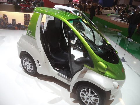 Green and white single-seat microcar with open sides and a small rear box on a bright white show floor. Opens a larger view