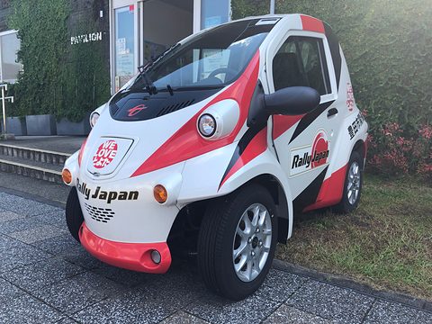 White single-seat microcar with red and black Rally Japan graphics parked on paving beside a hedge. Opens a larger view