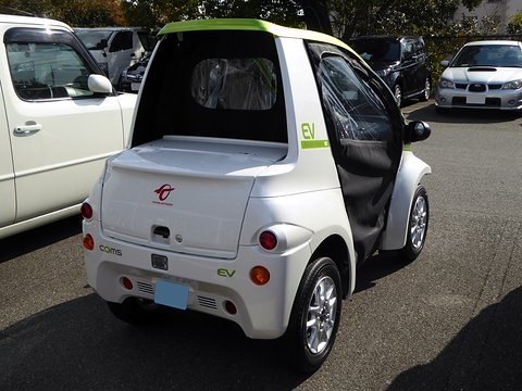 Rear of a white single-seat microcar with a small lockable boot box and round tail lamps in a car park. Opens a larger view