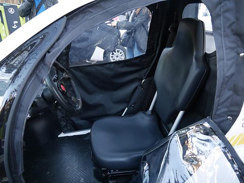 Black single seat with a grab bar and rubber floor mat inside a small car with a rolled-back clear door curtain. Opens a larger view