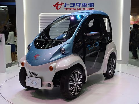 Pale blue and white single-seat microcar with a T-COM plate on a white stand under a Toyota Auto Body sign. Opens a larger view
