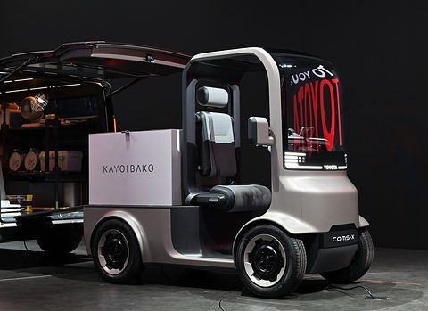 Grey open single-seat concept with a cargo box marked KAYOIBAKO and a lit screen, on a dark stand. Opens a larger view