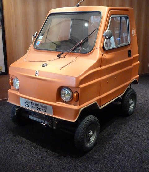 Orange boxy four-wheeled microcar with a roof aerial and a card reading Les petits ruisseaux 23 juin 2010 on its bumper.