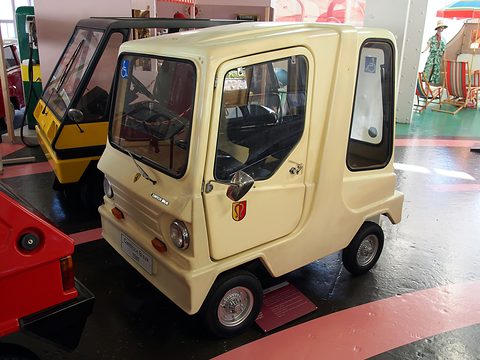 Cream tall box-shaped microcar with a large glasshouse and a wheelchair sticker, on a dark floor with pink stripes.