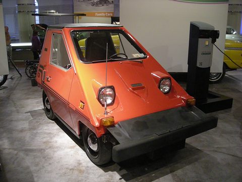 Orange-red wedge-shaped electric car with a deep black front bumper and a radio aerial, beside a charging post on a museum floor. Opens a larger view