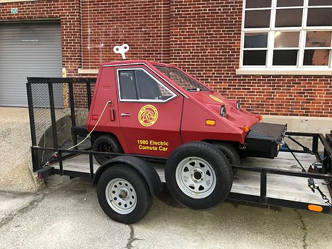 Maroon wedge-shaped electric car with a gold emblem and lettering on its door, on an open mesh trailer in front of a brick building. Opens a larger view