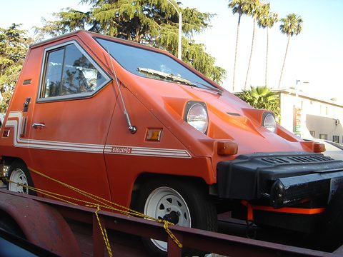 Orange wedge-shaped electric car with white side stripes and a deep black front bumper, strapped onto a trailer under palm trees. Opens a larger view