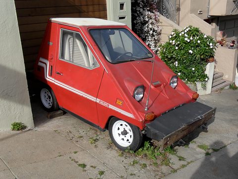 Red wedge-shaped electric car with a white roof, white side stripes and a deep black front bumper, parked by a flowering shrub and a garage. Opens a larger view