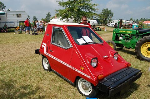 Red wedge-shaped electric car with white stripes and a black bumper box, parked on dry grass with a green and yellow tractor behind. Opens a larger view