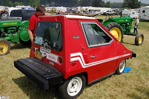 Rear quarter of a red wedge-shaped electric car with a large rear window, white side stripes and a black rear bumper box, tractors behind. Opens a larger view