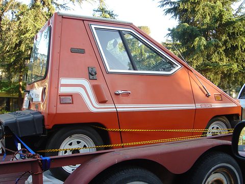 Side view of an orange wedge-shaped electric car with striped flanks, a door badge and a black battery-box bumper, tied down on a trailer. Opens a larger view