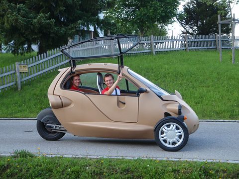 Beige three-wheeled two-seater with an open canopy frame and tandem seats on a lane beside a wooden fence.