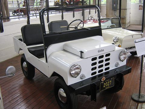 Small white open utility with a cross-hatch grille, round headlamps and a Crofton badge on its bonnet, on a wooden museum floor. Opens a larger view