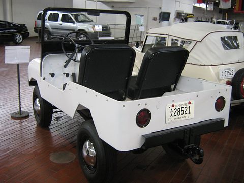 Rear quarter of a small white open utility with bucket seats, a roll hoop and a short load area, on a wooden museum floor.