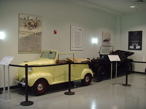 Small pale yellow open two-seater with its top folded, roped off in a bright museum gallery with framed posters. Opens a larger view