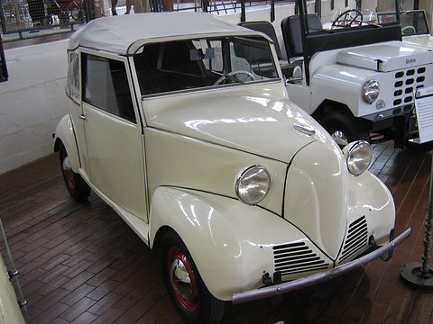 Small white two-door with a pale fabric top, V-shaped nose and louvred valance, on a brick museum floor. Opens a larger view