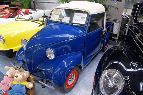 Small blue pick-up with a white canvas cab top and red wheels, teddy bears on display beside it.