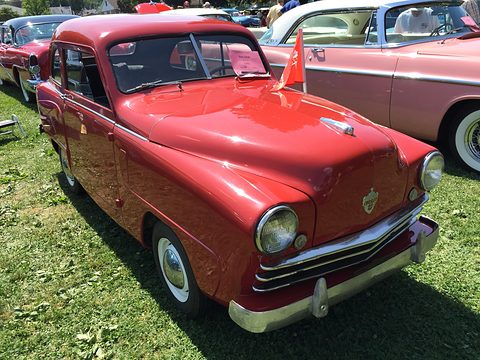 Red small saloon with a chrome grille and whitewall tyres parked on grass among other cars. Opens a larger view