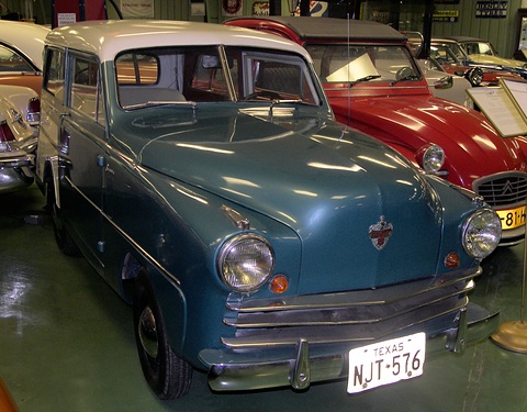 Teal small estate with a chrome grille and a Texas plate in a museum line-up beside a red car. Opens a larger view