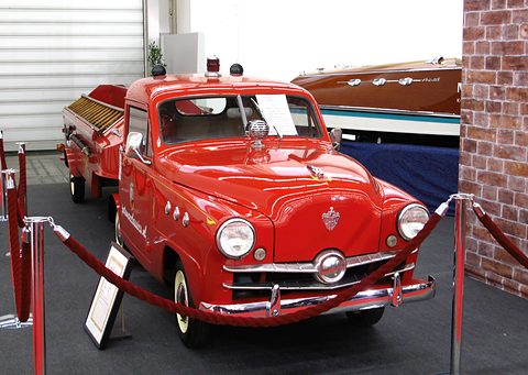 Red small pick-up with fire-service fittings behind a rope barrier, a wooden speedboat displayed behind. Opens a larger view