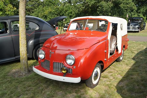 Red small open utility with a white soft top and no doors, parked on grass by a tree. Opens a larger view