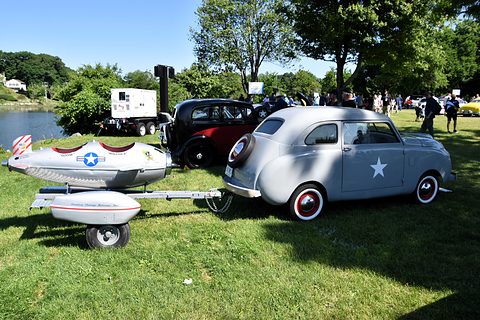Silver small saloon with white star roundels on its doors, towing a bullet-shaped trailer across a lawn. Opens a larger view