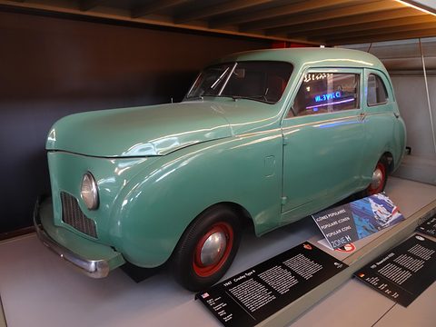 Turquoise slab-sided small saloon with red wheels in a display bay, information panels in front. Opens a larger view