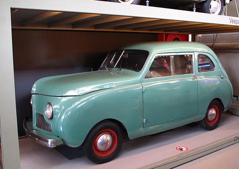 Side view of a turquoise slab-sided small saloon with red wheels on a white plinth. Opens a larger view