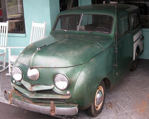 Weathered green small estate with twin round headlamps parked on a porch beside white rocking chairs. Opens a larger view