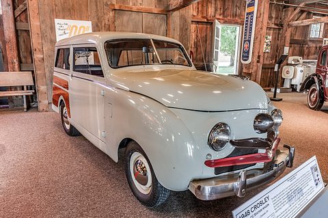 Grey small estate with woodgrain sides and a bull-nose grille inside a timber barn, a placard beside it. Opens a larger view