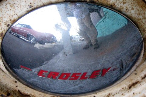 Rusty chrome hubcap with red CROSLEY lettering, reflecting a red car and the photographer. Opens a larger view