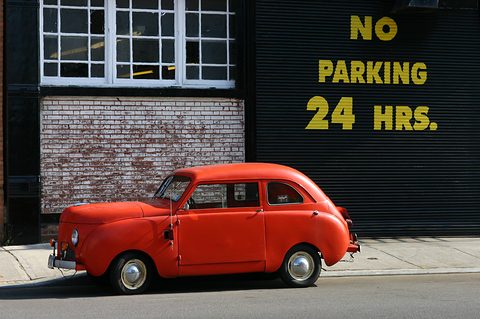 Bright red small saloon at the kerb beneath a sign reading NO PARKING 24 HRS on a dark shutter. Opens a larger view