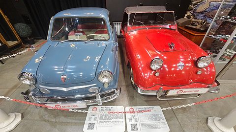Pale blue small saloon and red roadster parked together behind a chain, information boards in front. Opens a larger view