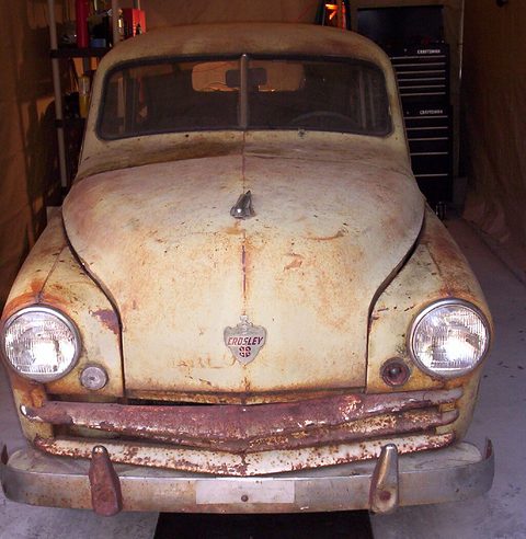 Heavily rusted small saloon seen head-on in a garage, its round headlamps and crest badge intact. Opens a larger view