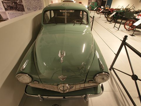 Front of a sage-green small saloon with a chrome grille and a bird mascot, display stands beside it. Opens a larger view