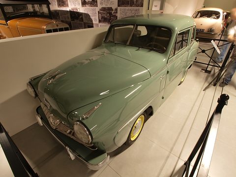 Sage-green small saloon with yellow wheels, seen from above the left front through a wide-angle lens. Opens a larger view