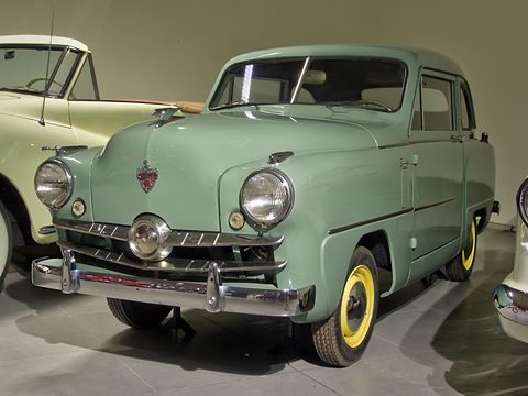 Sage-green small saloon with yellow wheels and a chrome grille, seen close from the right front. Opens a larger view
