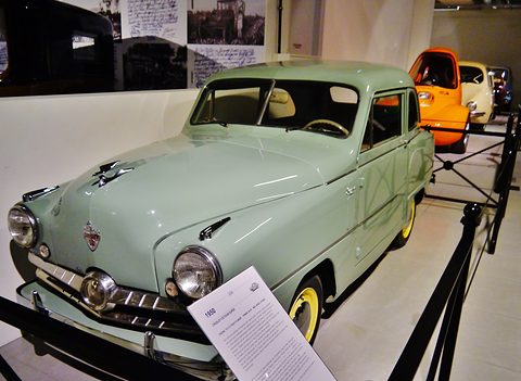 Sage-green small saloon with an information card on a stand in front, an orange car behind. Opens a larger view
