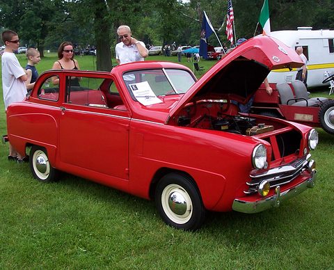 Red small two-door saloon with its bonnet open on a lawn, visitors and flags behind. Opens a larger view