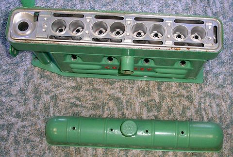 Green-painted engine block from above with eight valve openings, its cam cover lying below on carpet. Opens a larger view