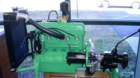 Green four-cylinder engine with radiator, starter and gearbox mounted on a wooden stand by a window. Opens a larger view