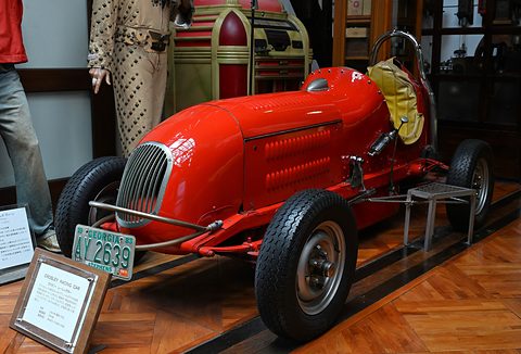 Red open-wheeled single-seat racing car with a vertical-slat grille and a plate, mannequins behind. Opens a larger view