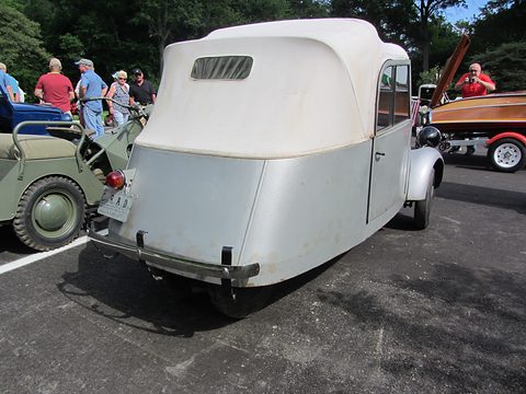 Silver-grey tiny car with a tall white fabric top and a tapering tail on closely set rear wheels, show visitors behind.