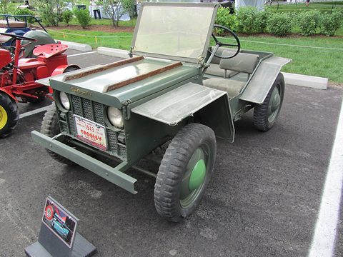 Tiny olive-green open 4x4 with a flat windscreen and square wings, parked on tarmac beside a display sign. Opens a larger view