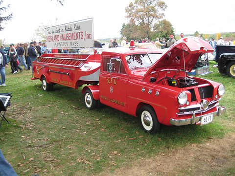 Red small fire truck with ladders and its bonnet open on a show field, a crowd and fairground sign behind. Opens a larger view