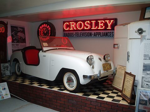 White doorless roadster on a chequered floor below a red neon sign reading CROSLEY RADIOS-TELEVISION-APPLIANCES.